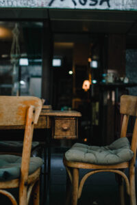 strassenszene-vor-einem-cafe-detailaufnahme-von-einem-holztisch-mit-schublade-davor-zwei-stuehle-auf-denen-sitzkissen-liegen-im-hinterfrund-verschwommen-der-eingang-des-cafes