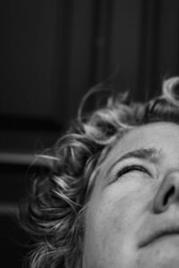 detailaufnahme-schwarzweiss-linke-gesichtshaelfte-einer-frau-mit-blonden-kurzen-locjen-sie-schaut-nach-oben-hat-das-auge-halb-verschlossen-von-der-sonne-geblendet