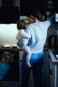 Eine Frau in einem hellen Strickpullover und Jeans steht mit dem Rücken zur Kamera in der Küche. Sie trägt ein Kleinkind auf dem Arm und greift in einen Oberschrank, während ein zweites Kind danebensteht.
