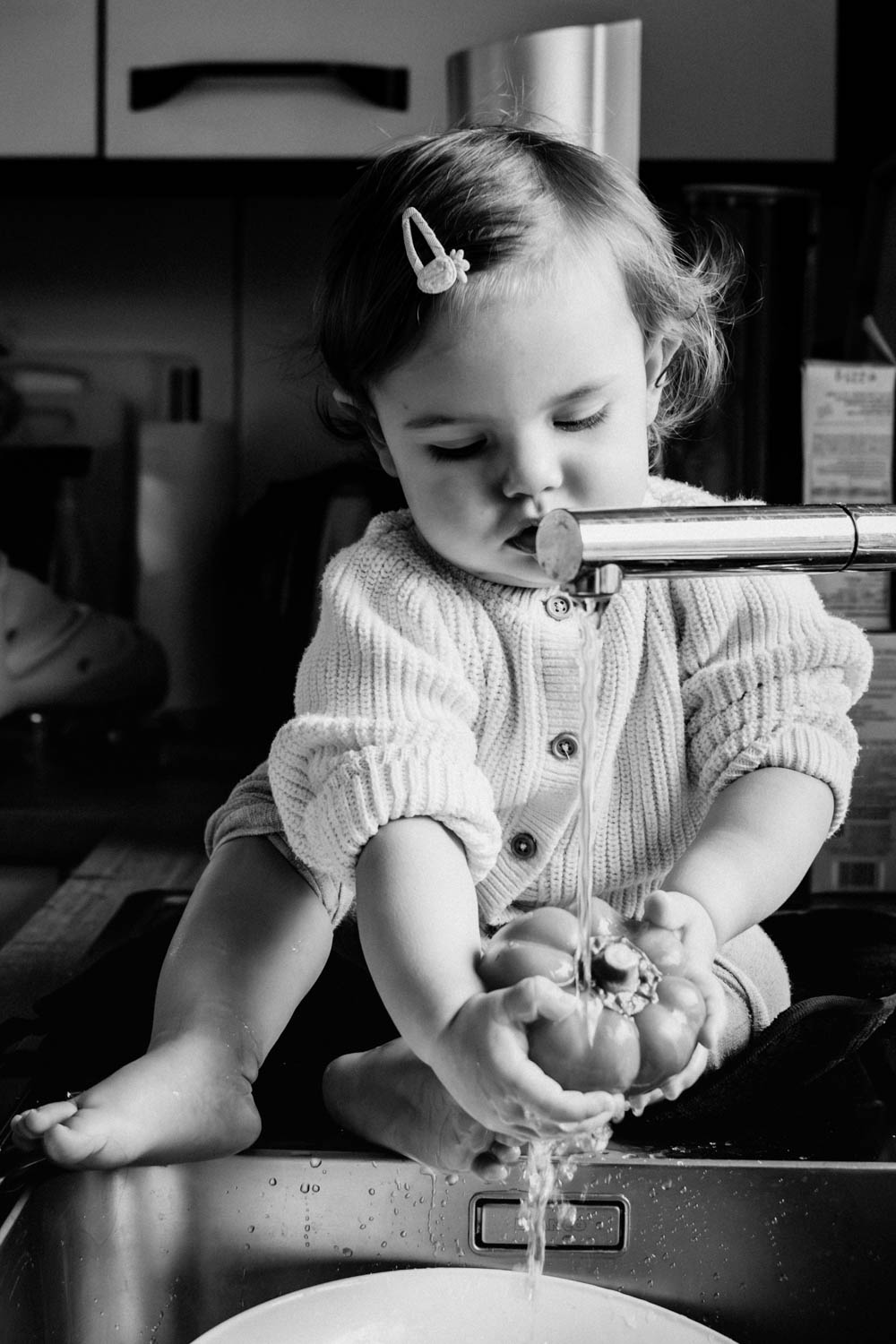 Schwarz-Weiß-Nahaufnahme eines Kleinkindes, das im Spülbecken sitzt und konzentriert eine Paprika unter fließendem Wasser wäscht.