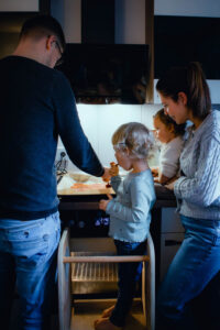 Eine Familie steht in der Küche an der Arbeitsplatte. Der Mann verteilt Tomatensauce auf einem Pizzateig, während die Frau und die beiden Kinder zuschauen. Ein Kind steht auf einem Lernturm.