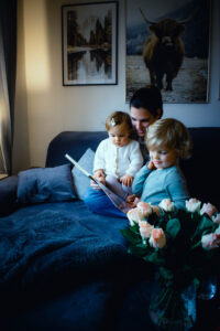 Eine Frau sitzt mit zwei kleinen Kindern auf einem dunklen Sofa und liest ihnen aus einem Buch vor. Im Vordergrund steht ein Strauß rosa Rosen, im Hintergrund hängen zwei Naturfotografien an der Wand.